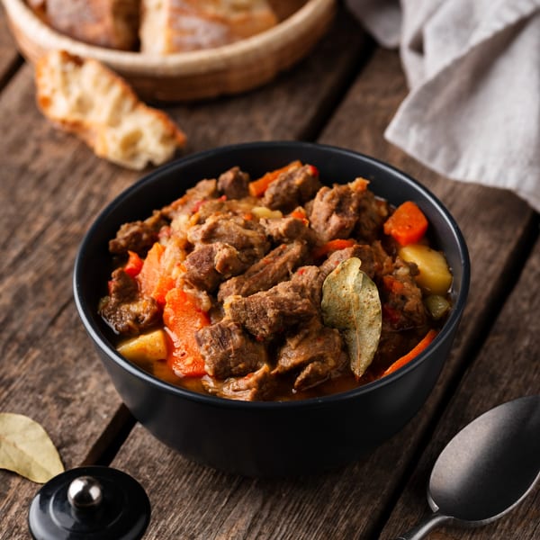 A black bowl of Indian beef stew with carrots and potatoes on a rustic wooden table.
